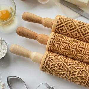 May include: Three wooden rolling pins with different engraved patterns. The patterns include a geometric design, a leaf design, and a diamond pattern. The rolling pins are made of natural wood and have a light brown color.