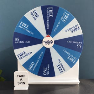 May include: A circular prize wheel with alternating dark and light blue segments, mounted on a white stand. The center features a logo reading The Laundry Spot. Text on segments includes Free Wash & Fold, Free Laundry Lab Bottle, Free Dryer Sheets, $5 Laundry Card, Free Add-on Service, $100 Laundromat Credits, One Free Wash, and Free Laundry Bag. A small white sign reads Take A Spin.