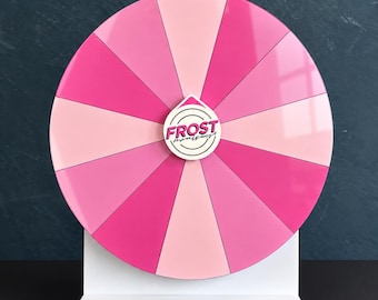 May include: A circular prize wheel featuring alternating segments of light pink and dark pink. The center of the wheel displays a white teardrop-shaped emblem with the text FROST written in bold pink letters and smaller cursive text below it.