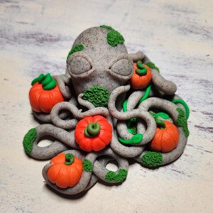 Puede incluir: Una escultura de arcilla gris y verde de un pulpo con cabeza de calabaza. El pulpo tiene ocho tentáculos y está decorado con pequeñas hojas verdes y cinco calabazas en miniatura.