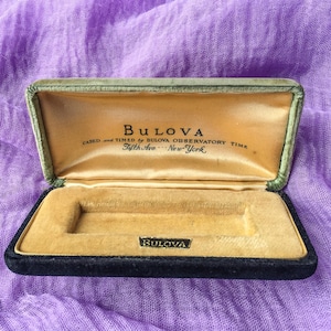 May include: Open vintage Bulova watch box. The box is black with a green velvet trim and a gold satin interior. The Bulova logo is visible on the inside of the lid and on a small metal plate inside the box. Text on the lid reads "Bulova Cased and Timed by Bulova Observatory Time Fifth Ave New York."