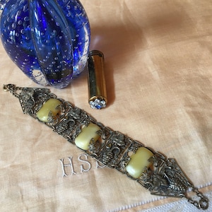 May include: An antique silver-tone bracelet with four rectangular yellow stones and grape vine detailing. A gold lipstick tube with a blue gem and a blue glass orb are also in the image. The initials "HS" are embroidered on the fabric.