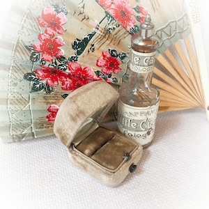 May include: An open, vintage, tan velvet ring box with two slots, next to a small glass cologne bottle with a silver cap. The bottle has a label that reads "Tappan's Little Cas Cologne". A floral fan is in the background.