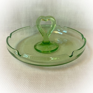 May include: A round, light green glass ashtray featuring a central handle shaped like a heart. The circular tray has a smooth, translucent finish with integrated notches along the outer rim, providing a functional design for resting items.