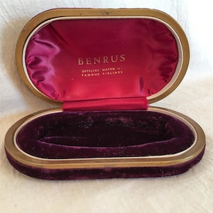 May include: An oval-shaped, gold-colored watch box with a burgundy velvet interior. The lid is open and the interior is lined with red satin. The lid has the words "BENRUS OFFICIAL WATCH U.S. FAMOUS AIRLINES" printed in gold lettering.