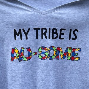 May include: A light gray V-neck t-shirt with the text "MY TRIBE IS AU-SOME". The word "AU-SOME" is in colorful puzzle piece letters. The shirt is held up by a person.
