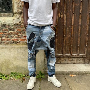May include: A person wearing a white polo shirt and blue denim jeans with patchwork details. The jeans have a unique design with different shades of blue and black denim sewn together.