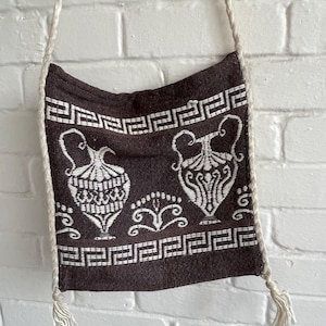 May include: A brown shoulder bag featuring a white Greek key design and vase illustrations. It has a cream rope strap with tassels. The bag is suspended against a white brick wall.