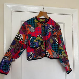 May include: A cropped jacket with a vibrant patchwork design. The jacket features a mix of patterns, including plaid, floral, and geometric shapes, in a palette of red, purple, orange, and black. It has a black trim and is hanging on a wooden hanger.