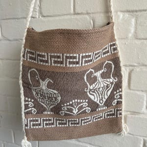 May include: A brown shoulder bag with white Greek key and vase designs. The bag has a white rope strap and is hanging against a white brick wall. The bag's design features a repeating pattern of white Greek keys.