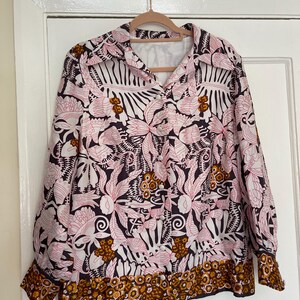 May include: A long-sleeved button-down shirt with a floral pattern in pink, white, brown, and gold. The shirt is hanging on a wooden hanger against a white door. The cuffs and hem have a decorative border.