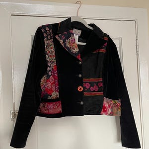 May include: Black velvet jacket with colourful floral patchwork panels. The jacket has a notched collar, three silver buttons, and a single orange button. The patchwork includes red, pink, and purple floral patterns. The jacket is hanging on a white hanger.