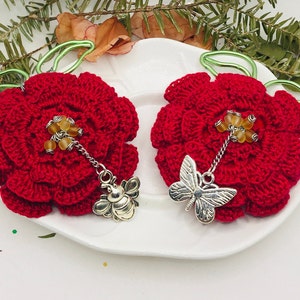 May include: Two handmade red crochet flower ornaments. Each flower has a silver charm, one a bee and the other a butterfly. The flowers have yellow bead accents and green wire hangers. These decorative ornaments are perfect for adding a touch of handmade charm.