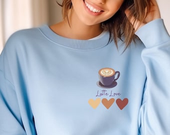 Puede incluir: Una sudadera de color azul claro con un gráfico de una taza de café con el texto "Latte Love" y tres corazones en diferentes tonos de marrón.