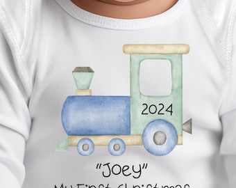 Pode incluir: Um macacão branco de manga comprida com um design de trem azul e verde. O trem tem o ano 2024 impresso nele. O texto "Joey" e "Meu primeiro Natal" está impresso abaixo do trem.