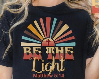 Puede incluir: Una camiseta negra con un gráfico de amanecer de estilo retro y el texto "Sé la luz" con el versículo bíblico "Mateo 5:14" debajo.