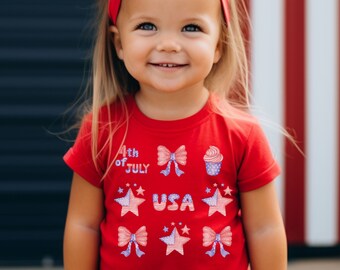 Puede incluir: Una niña pequeña con una camiseta roja con temática del 4 de julio. La camiseta tiene estrellas, lazos y una magdalena con detalles en rojo, blanco y azul. Lleva un lazo rojo, blanco y azul en el pelo.