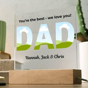 Op de afbeelding: Een helder acryl bord met het woord "DAD" in grote letters, met een landschapsontwerp in de letters. Het bord bevat de tekst "You're the best - we love you!" en de namen "Hannah, Jack & Chris". Het staat op een houten voet.