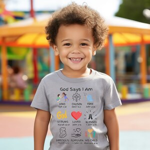 May include: A gray t-shirt with the text "God Says I Am" and illustrations of a cross, a bird, a heart, hands in prayer, a baby, and hands holding a heart. The text below each illustration includes a Bible verse.