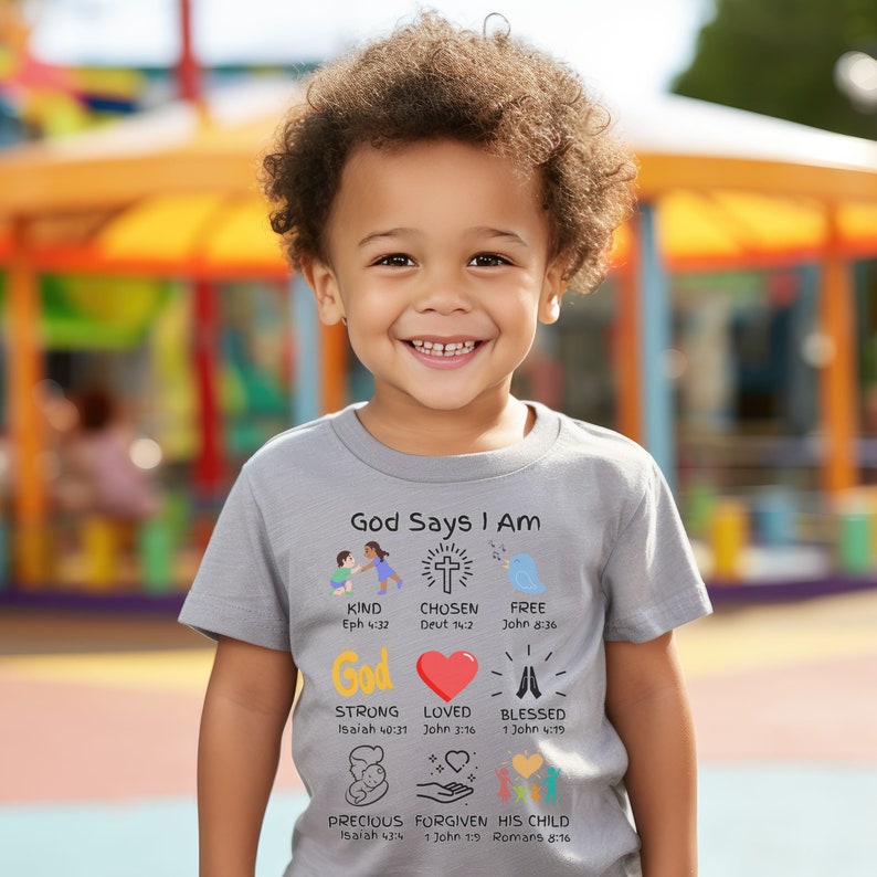 May include: A gray t-shirt with the text "God Says I Am" and illustrations of a cross, a bird, a heart, hands in prayer, a baby, and hands holding a heart. The text below each illustration includes a Bible verse.