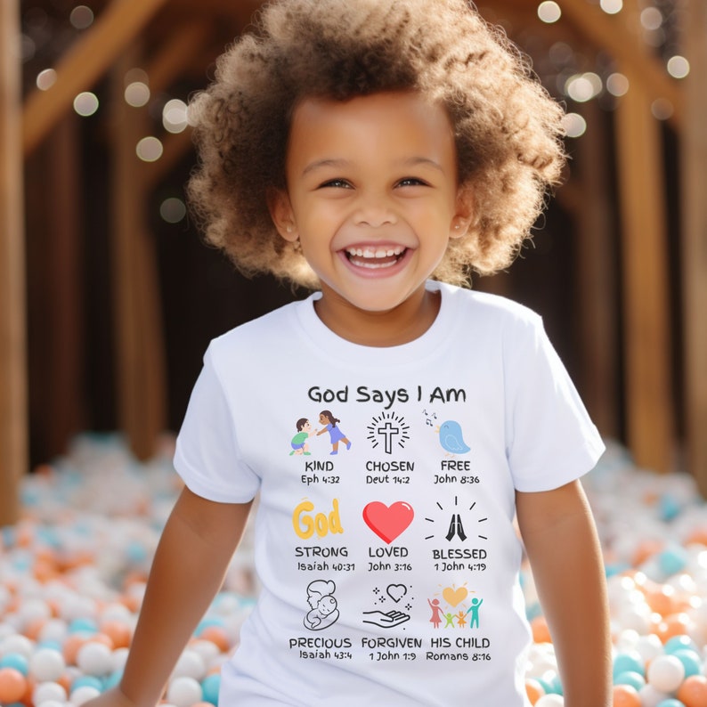 May include: A white t-shirt with the text 'God Says I Am' and illustrations of a cross, a heart, hands in prayer, and a baby. The text includes Bible verses for each illustration. The shirt is worn by a child with curly hair.