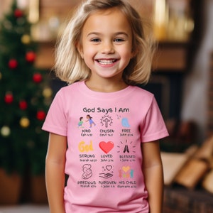 May include: Pink t-shirt with the text "God Says I Am" and illustrations of a cross, a heart, a bird, and hands. The text includes Bible verses for each illustration.