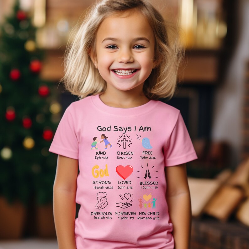 May include: Pink t-shirt with the text "God Says I Am" and illustrations of a cross, a heart, a bird, and hands. The text includes Bible verses for each illustration.