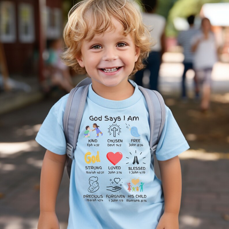 May include: A light blue t-shirt with the text "God Says I Am" and a variety of colorful illustrations and bible verses. The illustrations include a cross, a bird, a heart, hands in prayer, a baby, and hands holding a heart. The verses include "Eph 4:32", "Deut 14:2", "John 8:36", "Isaiah 40:31", "John 3:16", "1 John 4:19", "Isaiah 43:4", "1 John 1:9", and "Romans 8:16".