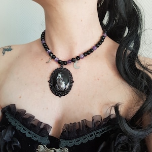 Peut inclure: Un collier ras de cou noir avec des perles violettes et un pendentif argenté représentant un camée d'une femme en robe noire. Le pendentif est entouré d'un cadre en filigrane noir.