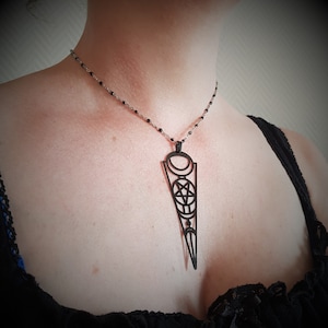 Op de afbeelding: Een zwarte hangerketting met een geometrisch ontwerp met een pentagram, halve manen en cirkels. De hanger hangt aan een fijne zilveren ketting met kleine zwarte kralen. De ketting wordt gedragen.