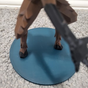 K&ouml;nnte beinhalten: Eine braune Figur mit schwarzen Akzenten steht auf einer t&uuml;rkisfarbenen, kreisf&ouml;rmigen Basis. Die Beine der Figur sind sichtbar, mit einem dunklen Objekt, das sich von der rechten Seite erstreckt. Die Basis befindet sich auf einem Teppichboden.