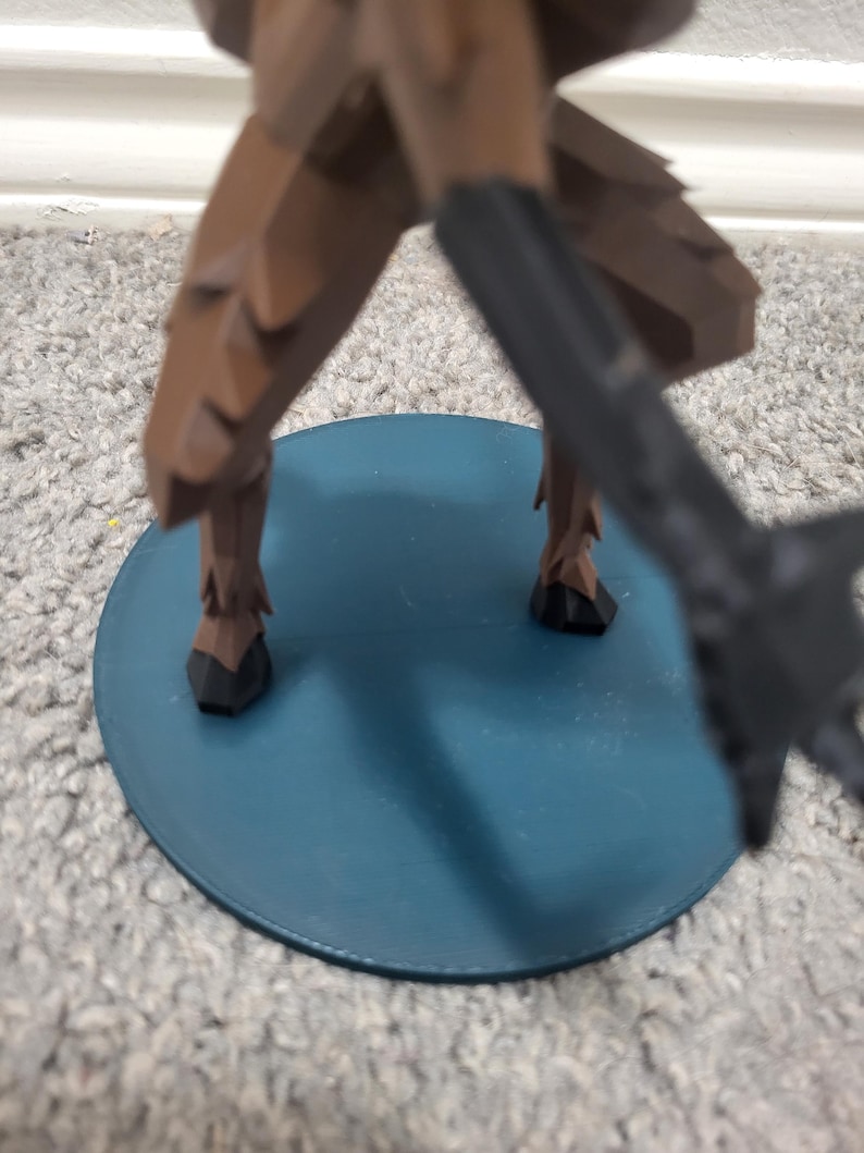 K&ouml;nnte beinhalten: Eine braune Figur mit schwarzen Akzenten steht auf einer t&uuml;rkisfarbenen, kreisf&ouml;rmigen Basis. Die Beine der Figur sind sichtbar, mit einem dunklen Objekt, das sich von der rechten Seite erstreckt. Die Basis befindet sich auf einem Teppichboden.