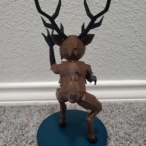 K&ouml;nnte beinhalten: Eine braune, geometrische Hirschfigur mit schwarzen Geweihen und Armen, die auf einer t&uuml;rkisfarbenen, kreisf&ouml;rmigen Basis posiert. Der Hirsch befindet sich in einer dynamischen Pose, mit einem erhobenen Arm. Die Figur ist wahrscheinlich 3D-gedruckt und hat ein einzigartiges, eckiges Design.