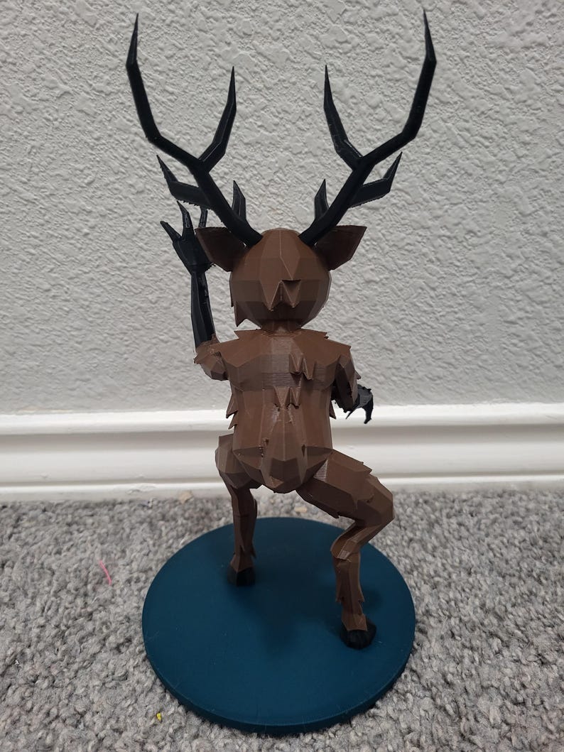 K&ouml;nnte beinhalten: Eine braune, geometrische Hirschfigur mit schwarzen Geweihen und Armen, die auf einer t&uuml;rkisfarbenen, kreisf&ouml;rmigen Basis posiert. Der Hirsch befindet sich in einer dynamischen Pose, mit einem erhobenen Arm. Die Figur ist wahrscheinlich 3D-gedruckt und hat ein einzigartiges, eckiges Design.