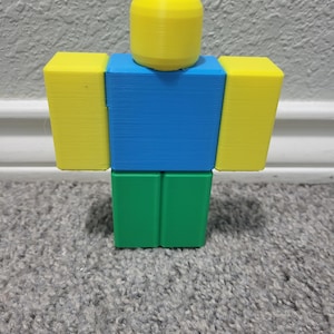 May include: A 3D-printed figure in the shape of a Roblox character. The figure has a yellow head and arms, a blue torso, and green legs. The figure is standing on a gray carpet.