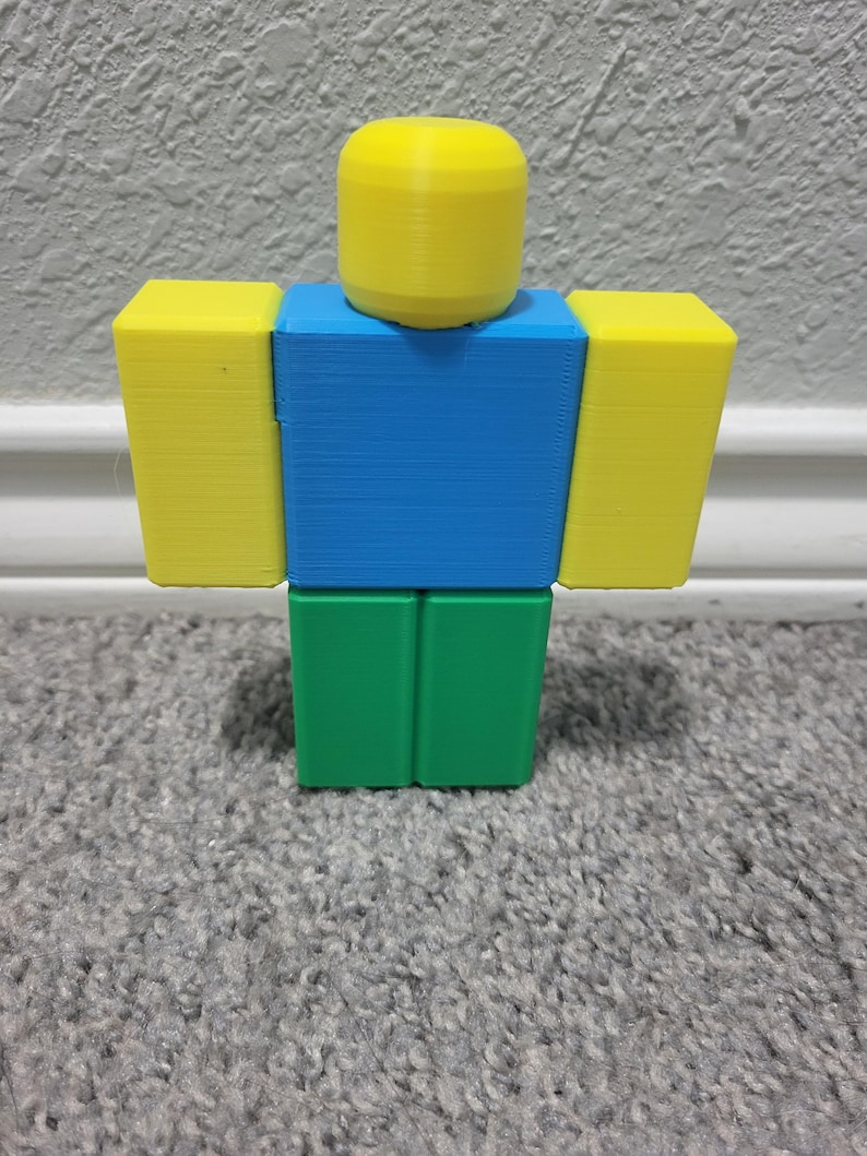 May include: A 3D-printed figure in the shape of a Roblox character. The figure has a yellow head and arms, a blue torso, and green legs. The figure is standing on a gray carpet.