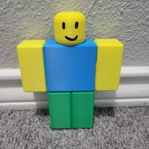 May include: A 3D-printed figure with a yellow block head featuring a smiling face. The figure has blue torso, green legs, and yellow block arms. The figure is standing upright.