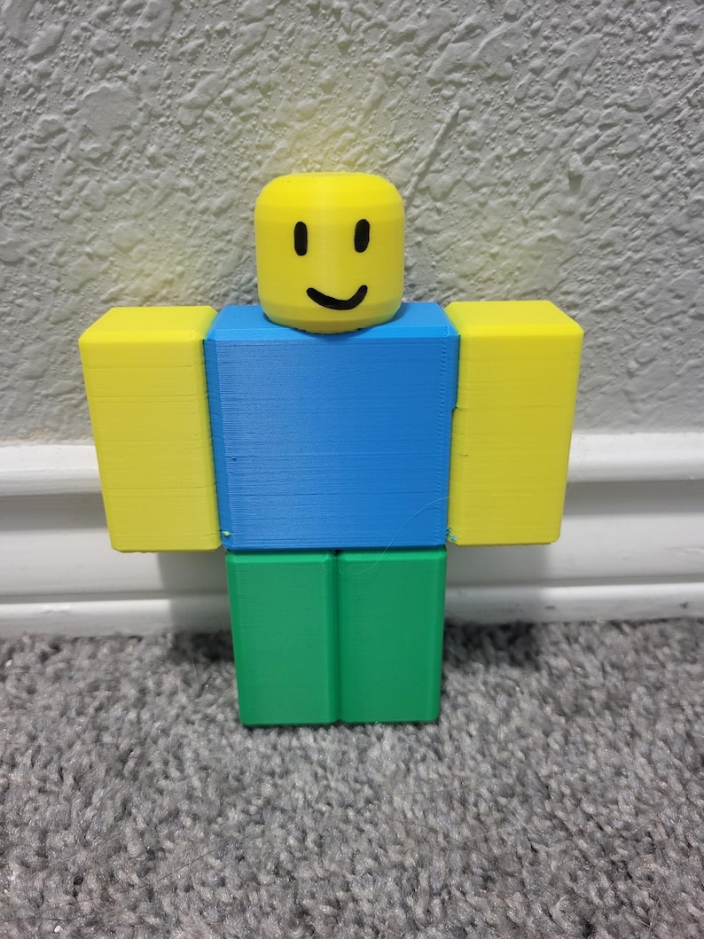 May include: A 3D-printed figure with a yellow block head featuring a smiling face. The figure has blue torso, green legs, and yellow block arms. The figure is standing upright.