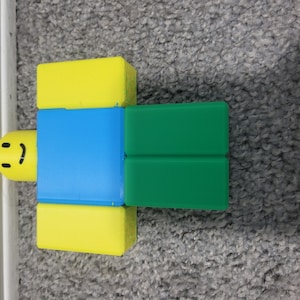May include: A colorful toy figure with a yellow head and legs, a blue torso, and green arms. The figure has a smiling face. The toy is designed in a blocky style, reminiscent of a popular video game character.