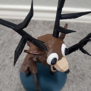 K&ouml;nnte beinhalten: Eine 3D-gedruckte Hirschfigur mit braunem K&ouml;rper, schwarzen Geweih und gro&szlig;en wei&szlig;en Augen. Der Hirsch hat ein geometrisches Design und ist auf einem blauen kreisf&ouml;rmigen Sockel montiert. Das Geweih ist spitz und kantig.