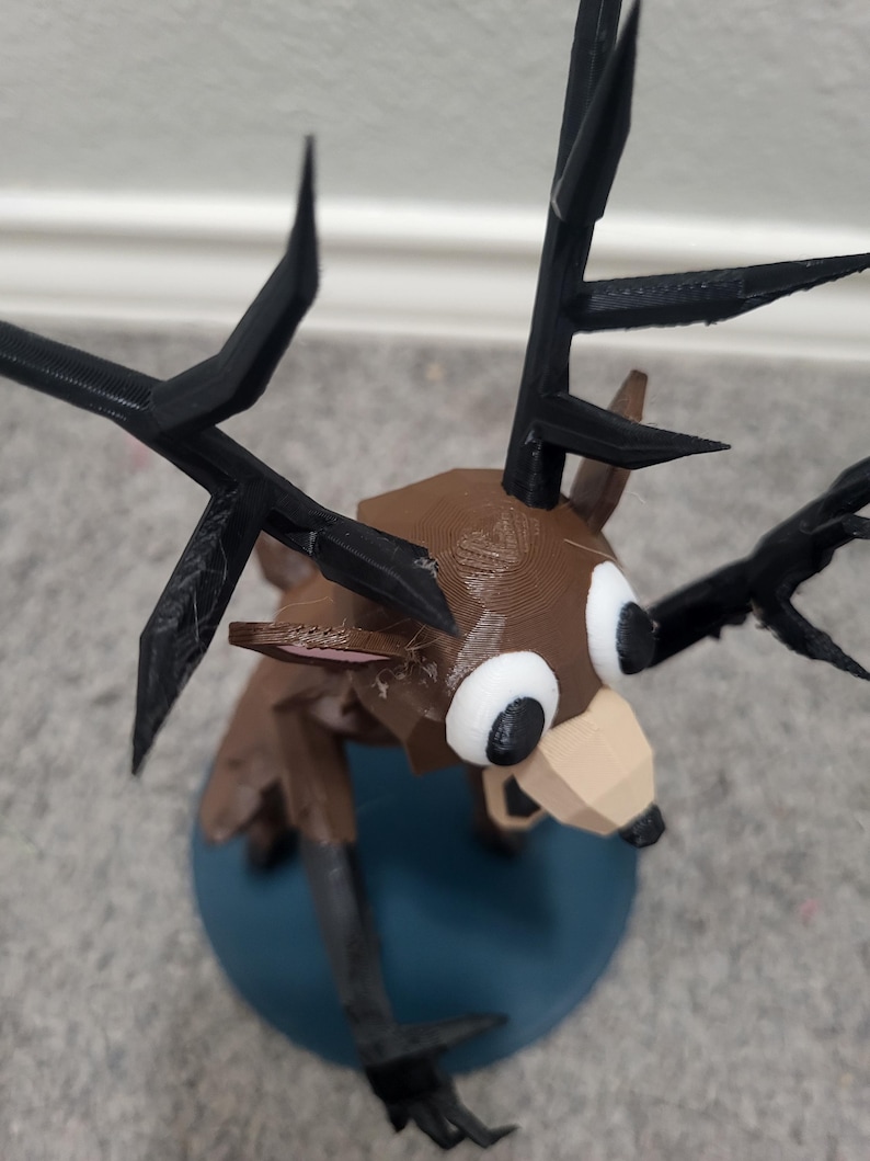K&ouml;nnte beinhalten: Eine 3D-gedruckte Hirschfigur mit braunem K&ouml;rper, schwarzen Geweih und gro&szlig;en wei&szlig;en Augen. Der Hirsch hat ein geometrisches Design und ist auf einem blauen kreisf&ouml;rmigen Sockel montiert. Das Geweih ist spitz und kantig.