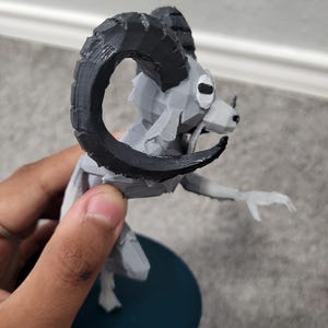 Op de afbeelding: Een 3D-geprint beeldje van een ramachtig wezen met grote, gebogen zwarte hoorns. De figuur is voornamelijk grijs met zwarte accenten en is gemonteerd op een donkerblauwe cirkelvormige basis. Het wezen heeft een gestileerd, geometrisch ontwerp.