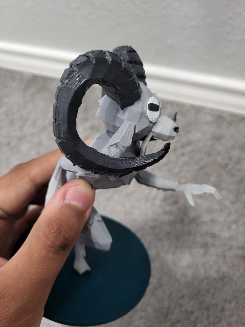 Op de afbeelding: Een 3D-geprint beeldje van een ramachtig wezen met grote, gebogen zwarte hoorns. De figuur is voornamelijk grijs met zwarte accenten en is gemonteerd op een donkerblauwe cirkelvormige basis. Het wezen heeft een gestileerd, geometrisch ontwerp.