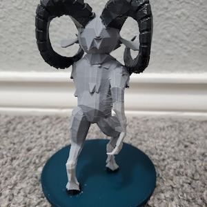 Op de afbeelding: Een grijze 3D-geprinte figuur van een mythisch wezen met grote, gebogen zwarte hoorns. De figuur heeft een geometrisch ontwerp en staat op een cirkelvormige, blauwgroene basis. De figuur staat op een grijs tapijt.