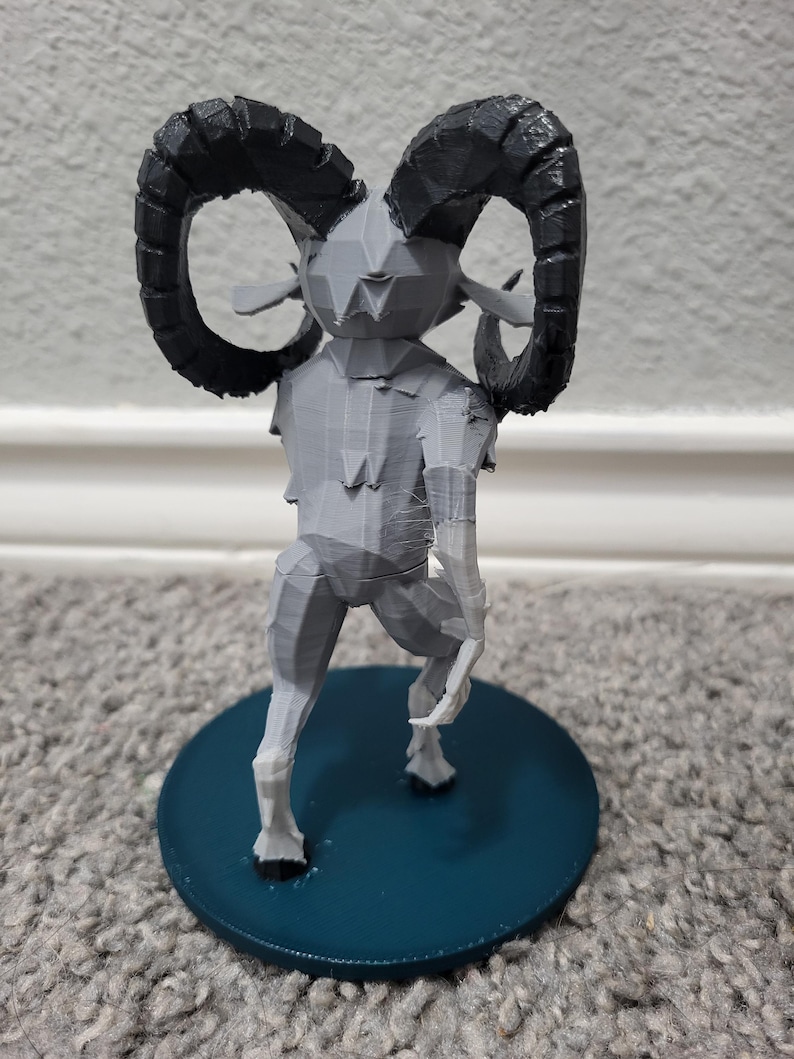 Op de afbeelding: Een grijze 3D-geprinte figuur van een mythisch wezen met grote, gebogen zwarte hoorns. De figuur heeft een geometrisch ontwerp en staat op een cirkelvormige, blauwgroene basis. De figuur staat op een grijs tapijt.