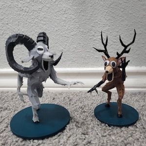 Op de afbeelding: Twee grillige beeldjes: een grijze ram met grote hoorns en een bruin hert met geweien. Beide staan op teal-kleurige, cirkelvormige bases, met overdreven kenmerken en open monden, waardoor ze een cartoonachtig uiterlijk hebben. De figuren staan tegen een neutrale achtergrond.