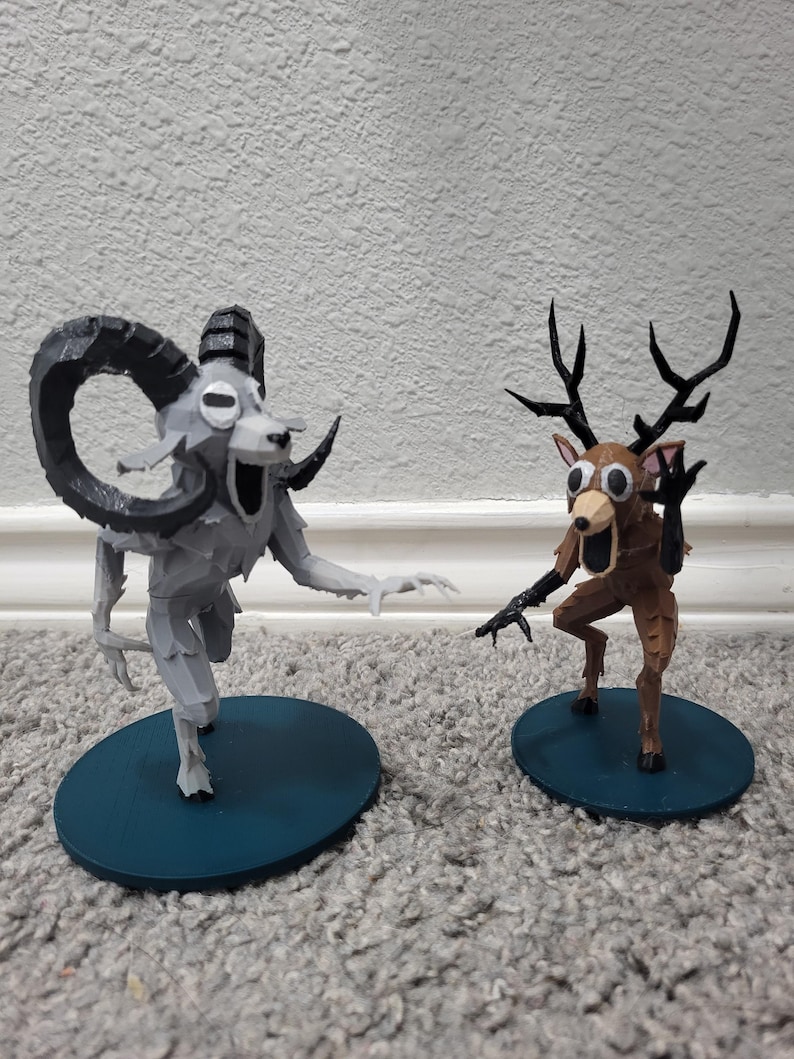 Op de afbeelding: Twee grillige beeldjes: een grijze ram met grote hoorns en een bruin hert met geweien. Beide staan op teal-kleurige, cirkelvormige bases, met overdreven kenmerken en open monden, waardoor ze een cartoonachtig uiterlijk hebben. De figuren staan tegen een neutrale achtergrond.