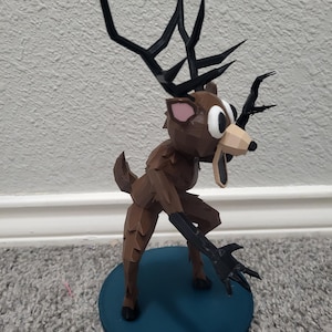 K&ouml;nnte beinhalten: Eine braune, geometrische Hirschfigur mit gro&szlig;en schwarzen Geweihen und schwarzen Krallen. Der Hirsch steht auf einem kreisf&ouml;rmigen Sockel in T&uuml;rkis. Die Augen des Hirsches sind gro&szlig; und wei&szlig; mit schwarzen Pupillen.
