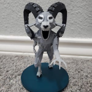 Op de afbeelding: Een 3D-geprinte figuur van een op een ram lijkend wezen met grote gebogen hoorns en een open mond. De figuur is voornamelijk grijs met zwarte hoorns en staat op een cirkelvormige teal-kleurige basis.