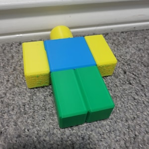 May include: A colorful, blocky figure with a yellow head and arms, a blue torso, and green legs. The figure is a popular character from a video game. The figure is resting on a gray carpet.